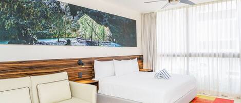 A hotel room with a bed, a sofa, a ceiling fan, and a large painting of a forest scene.
