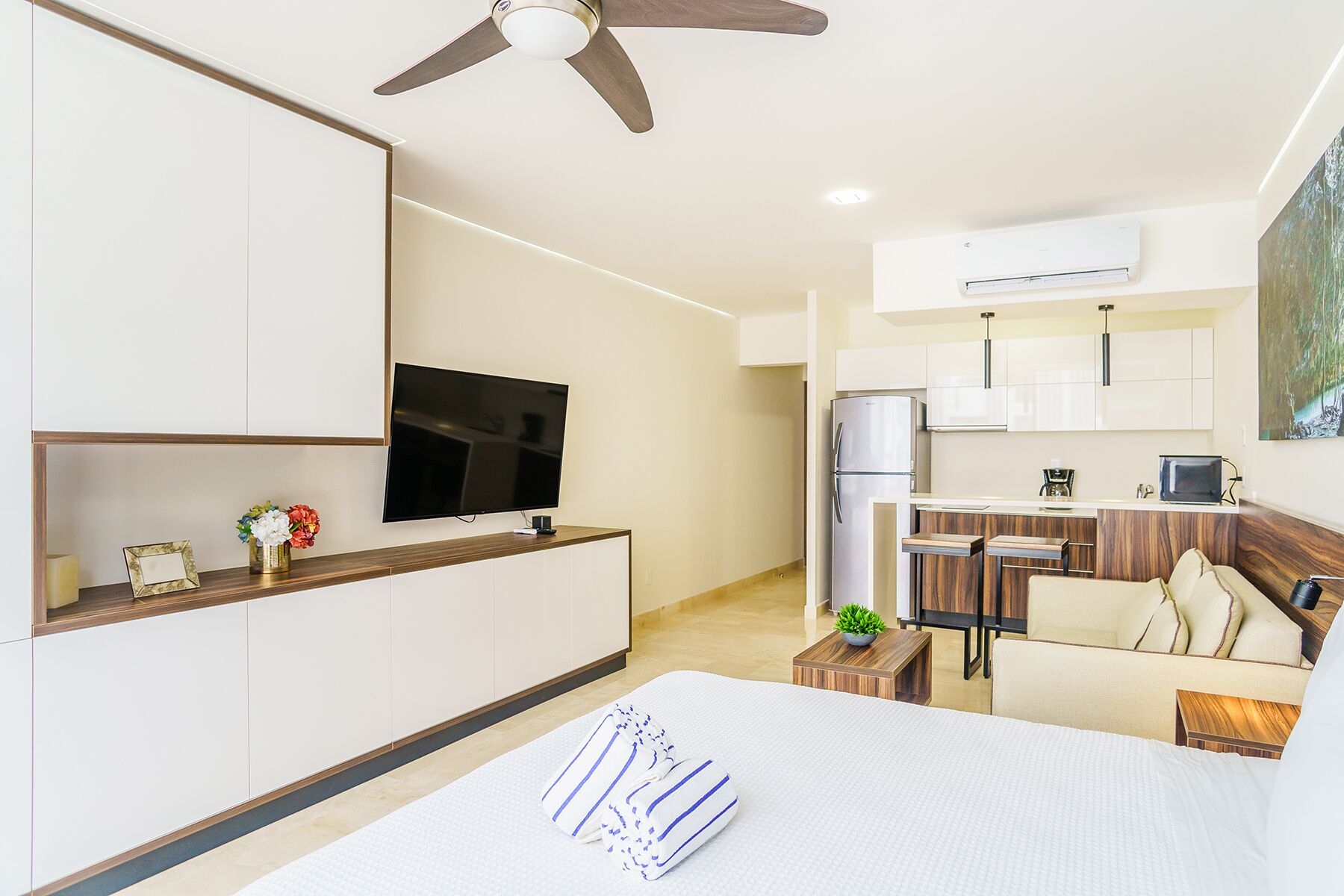 A modern hotel room with a bed, a TV, a kitchenette, and a ceiling fan.