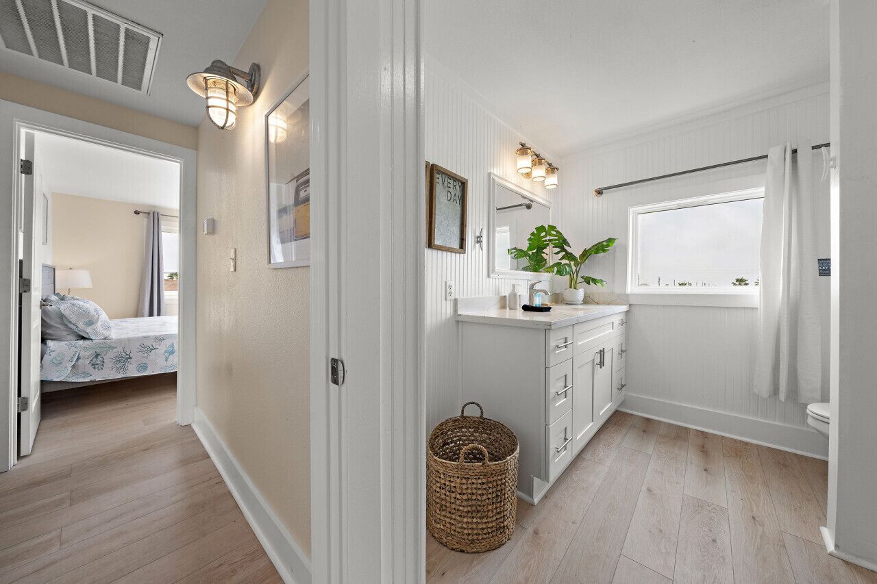 A modern bathroom with a shower, toilet, and sink. There is a window with a curtain, a plant on the counter, and a basket on the floor.