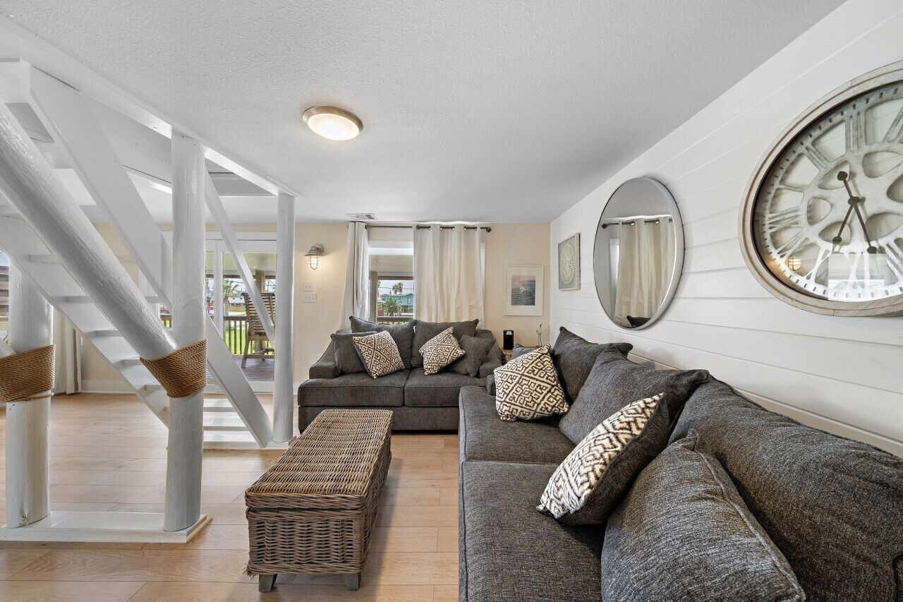 A modern living room with a grey sofa set, a wicker ottoman, and a large wall clock.
