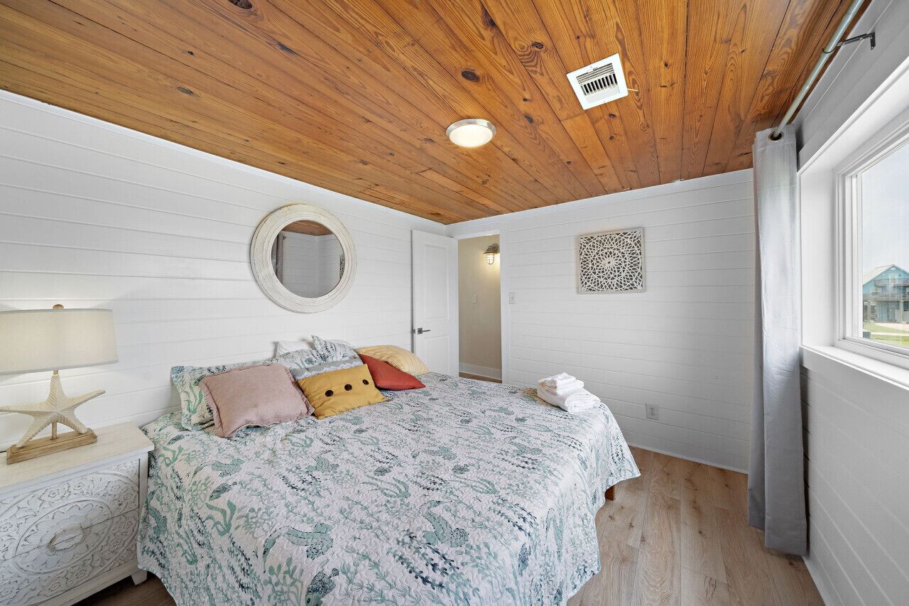 A bedroom with a wooden ceiling, a bed with patterned bedding, a round mirror, and a decorative wall piece.