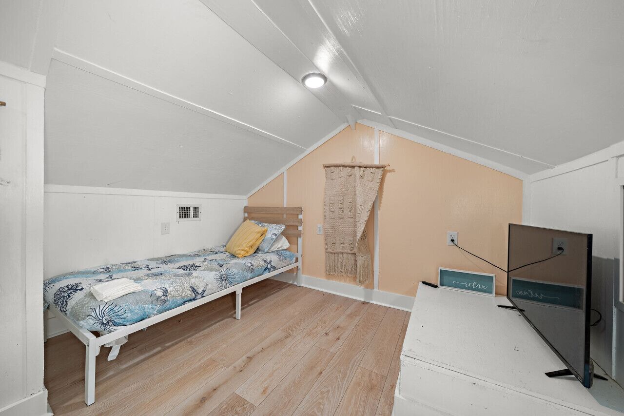 Un dormitorio pequeño y luminoso con una cama individual, suelo de madera, techo inclinado y un televisor sobre un mueble.
