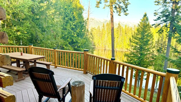 A wooden deck with picnic tables and chairs, surrounded by a railing, overlooking a forested area. - Montana Mountain Log-Lodge with lake views, lake access & mountain views (Bigfork)