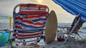 A beach chair with a striped beach bag and a surfboard, under a blue umbrella, with the ocean and sunset in the background.