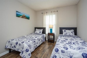 Una habitación con dos camas, un cuadro en la pared, una ventana con cortinas y una mesita de noche con lámpara.
