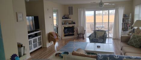 A spacious living room with a television, seating area, and a view of the beach through large windows.