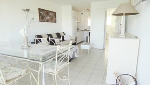 Ein modernes, helles Wohnzimmer mit einem Glastisch, weißen Sofas und einer Küche im Hintergrund.