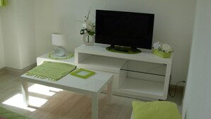 Een moderne, minimalistische kamer met een wit bureau, een televisie op een standaard, een klein wit kastje en een groene kussen op een witte stoel.