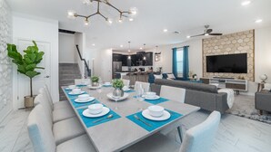 A modern dining area with a long table set for six, a kitchen in the background, and a living area with a TV and seating. - 5 Star Villa Zenith – The Summit of Opulent Living (Orlando)
