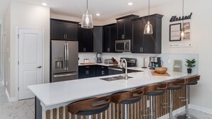 A modern kitchen with dark cabinets, stainless steel appliances, a white countertop, and wooden bar stools. - 5 Star Villa Zenith – The Summit of Opulent Living (Orlando)