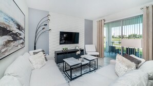 Een moderne woonkamer met een witte bank, een zwarte salontafel, een flatscreen televisie bevestigd aan een witte bakstenen muur en een balkon met uitzicht.
