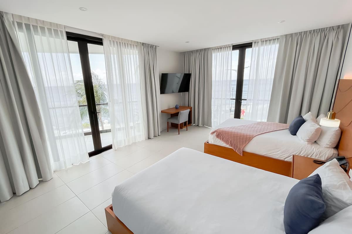 Quarto de hotel moderno com duas camas, uma mesa de trabalho, televisão e janelas amplas com cortinas.