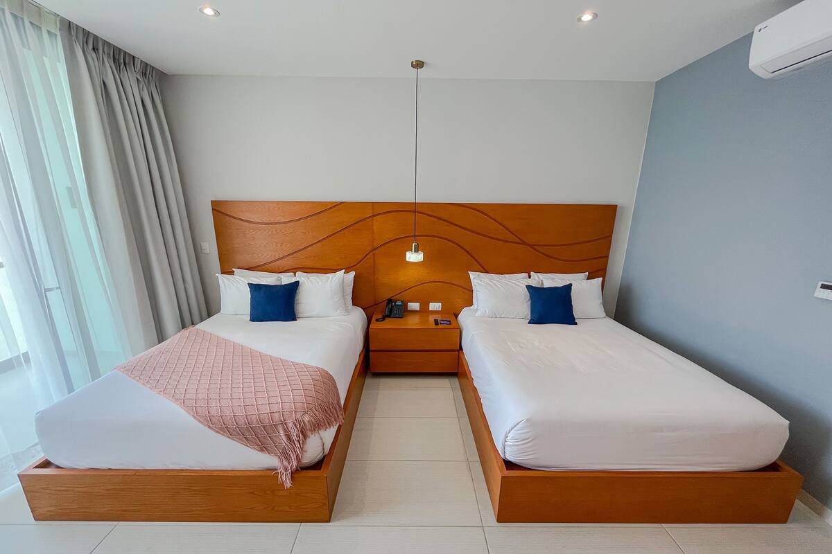 Quarto duplo com duas camas, cabeceira de madeira, mesa de cabeceira com luminária e janela com cortinas.