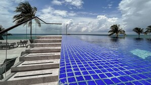 A poolside area with a blue tiled floor, steps leading to the water, palm trees, and a clear sky.