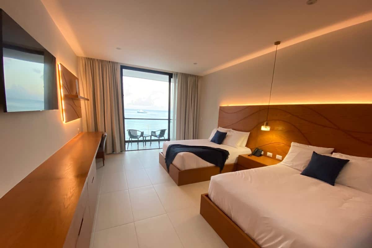 Quarto de hotel com duas camas, uma escrivaninha e varanda com vista para o mar.