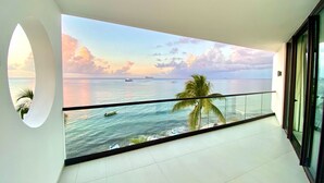 Un balcón con vista al océano y barcos, con una palmera y barandilla de cristal.