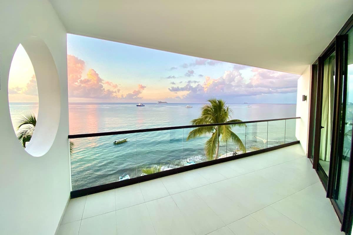 Uma varanda com vista para o oceano e navios, com uma palmeira e guarda-corpo de vidro.