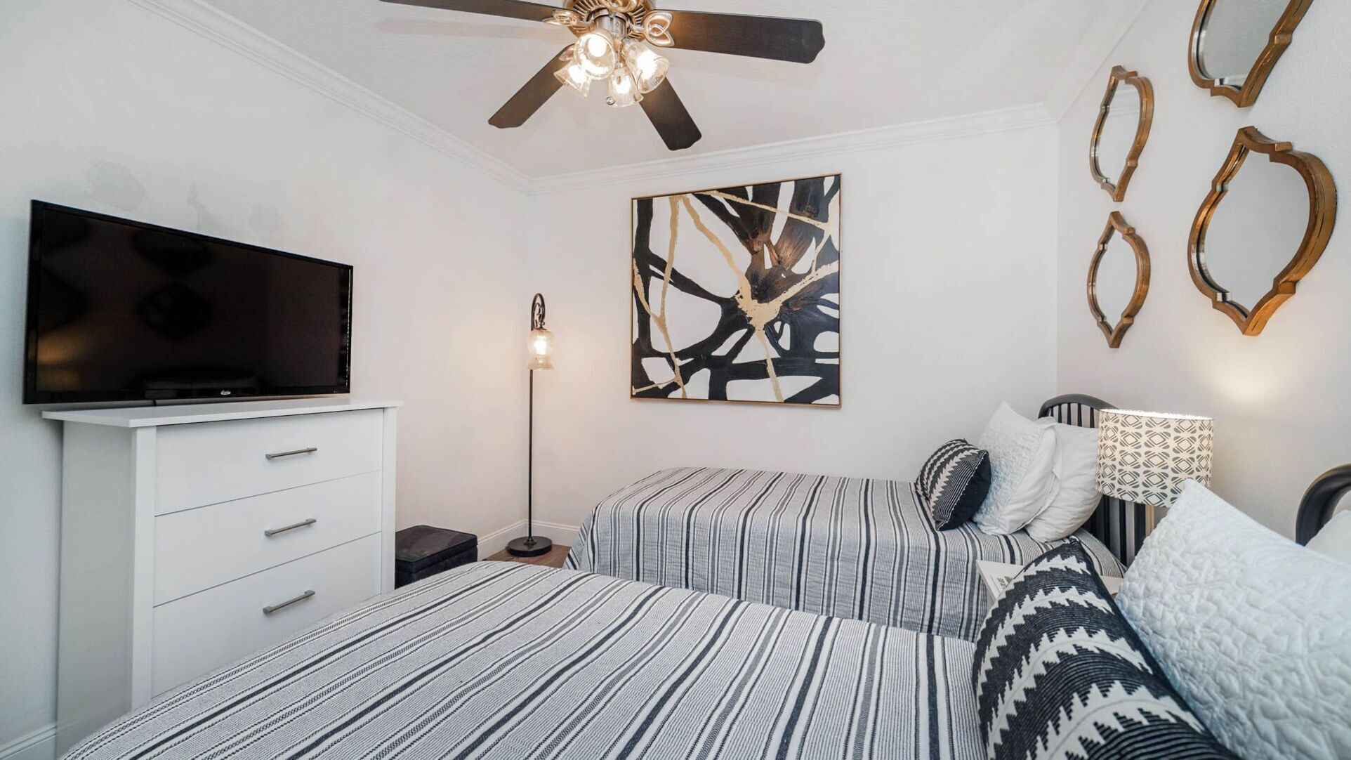 A bedroom with a TV on a white dresser, a ceiling fan, a floor lamp, two beds with striped bedding, a wall art piece, and decorative mirrors.