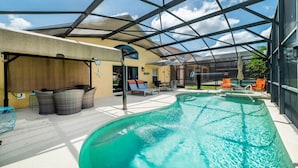 A covered swimming pool with lounge chairs and a sunbed, surrounded by a glass enclosure.