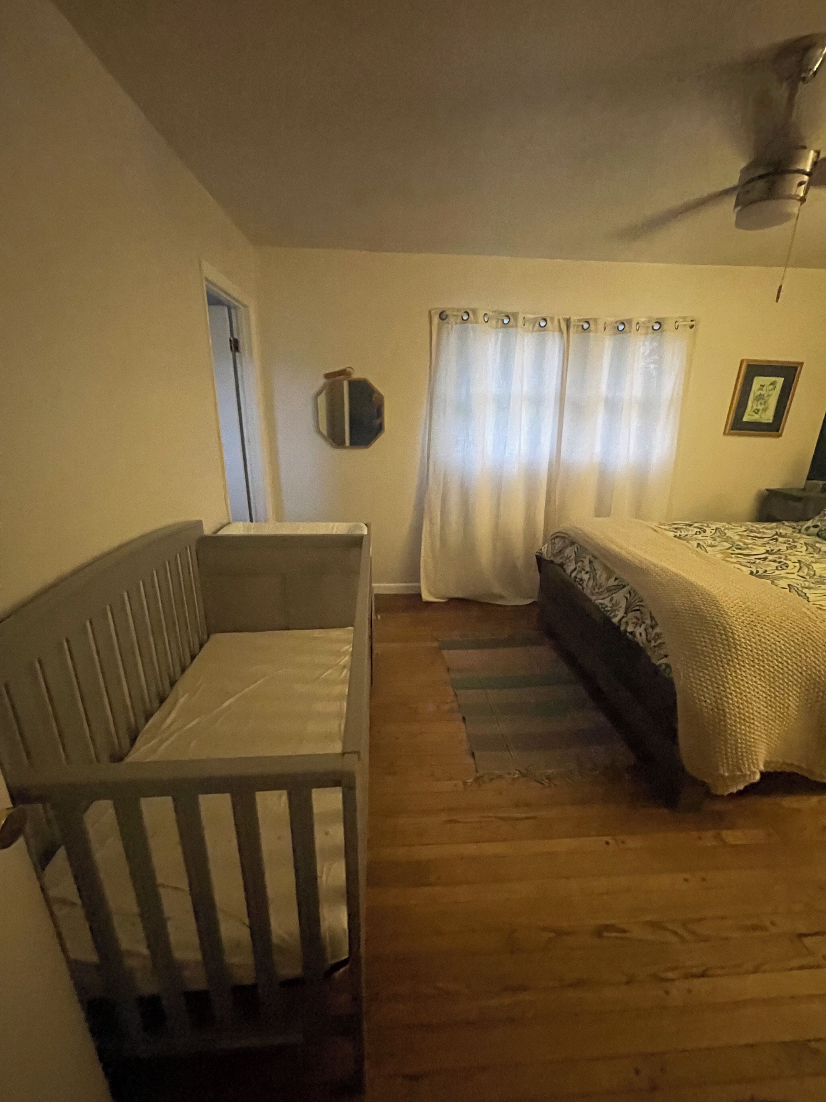 A room with a wooden floor, a bed with a patterned blanket, a crib, a ceiling fan, and a door.