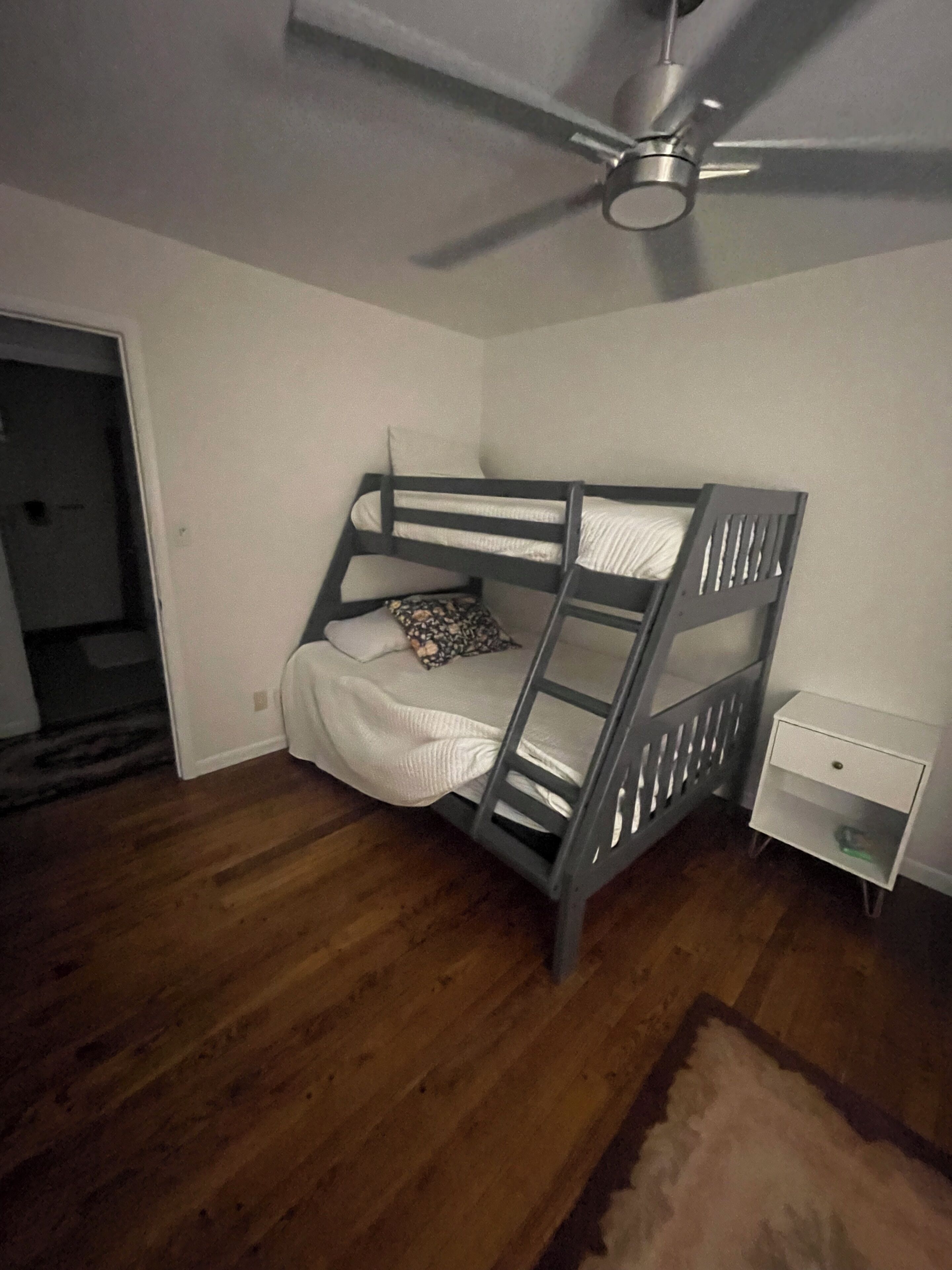 A room with a bunk bed, a white nightstand, a ceiling fan, and wooden flooring.