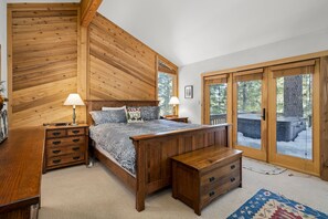 A bedroom with a wooden sloped ceiling, a large bed with a gray and white patterned bedspread, two bedside tables with lamps, a wooden bench, and a view through a glass door to the outside.