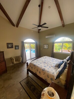 A bedroom with a bed, a ceiling fan, a dresser, and a window with a view of greenery.