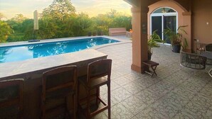 Zona de piscina con bar y asientos, rodeada por un porche cubierto con suelo estampado y plantas en macetas.