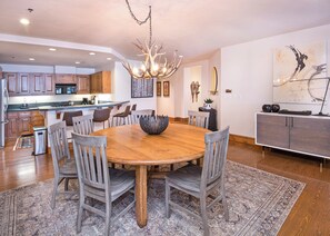 A spacious dining area with a round wooden table surrounded by chairs, a decorative chandelier, and a modern kitchen in the background. - Ski-in/out Beaver Creek condo, 4BR, 4.5BA, mountain views. (Beaver Creek)