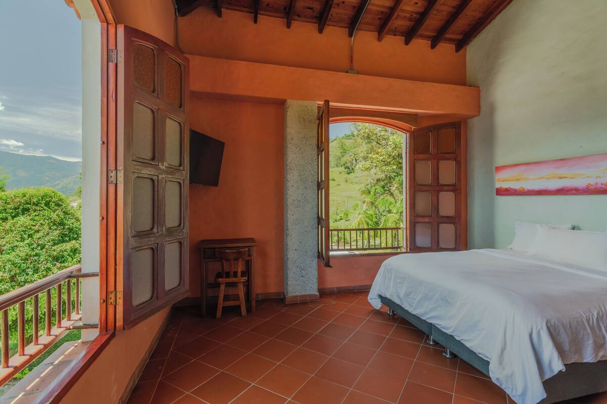 Quarto com cama, escrivaninha e cadeira, varanda com guarda-corpo e vista para o verde.