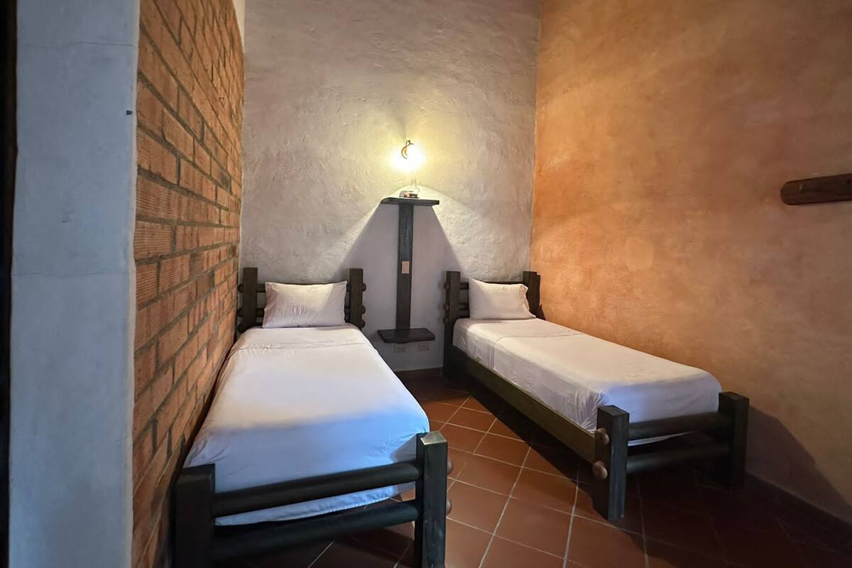 Quarto pequeno com duas camas de solteiro, parede de tijolos, chão de azulejos e luminária presa à parede.