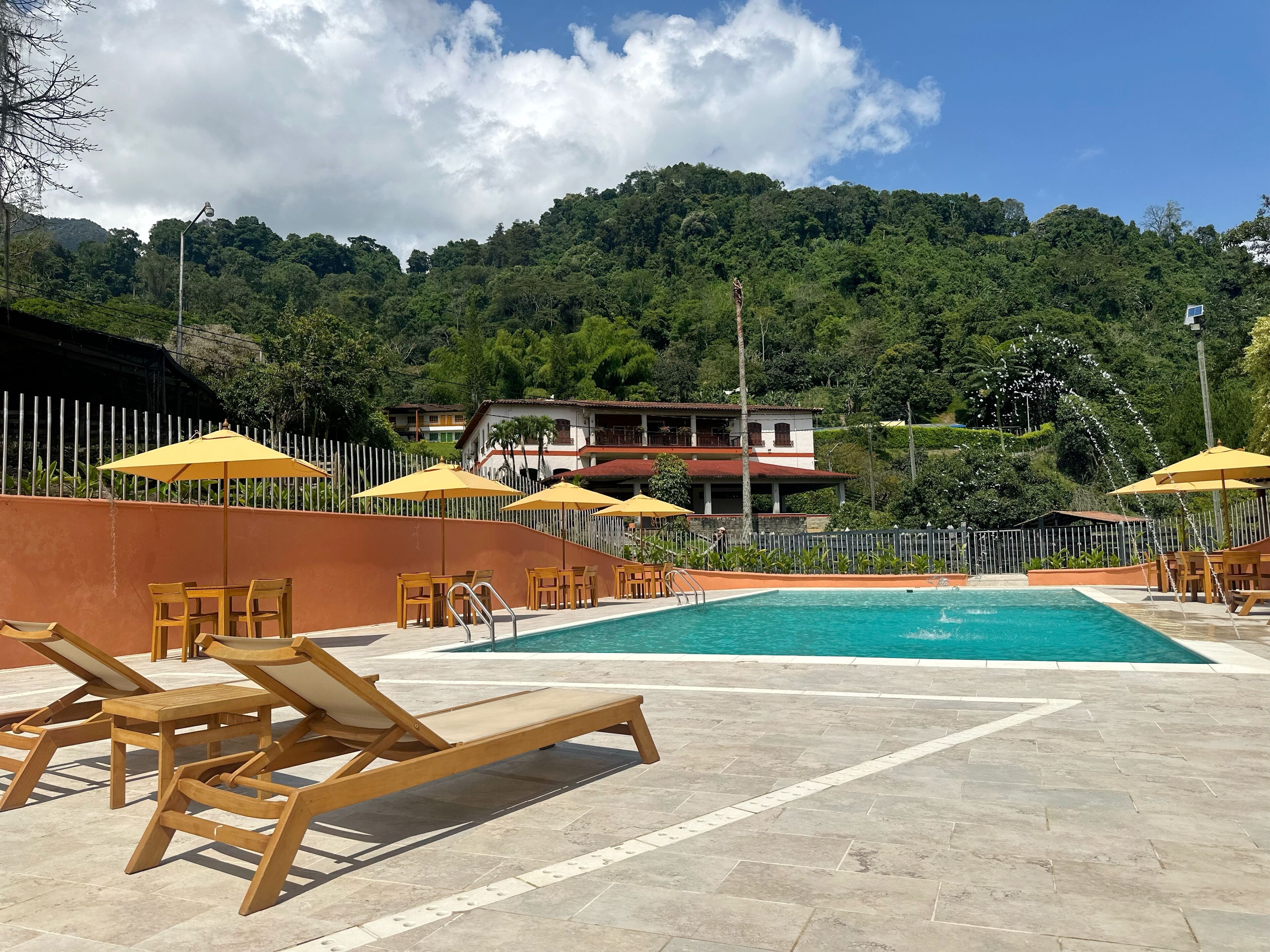 A pool area with wooden loungers and yellow umbrellas, surrounded by a white fence and a hillside backdrop.
