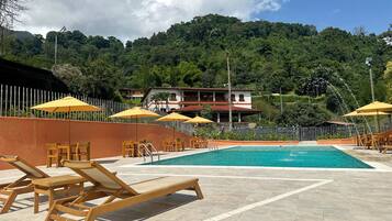 A pool area with wooden loungers and yellow umbrellas, surrounded by a white fence and a hillside backdrop.