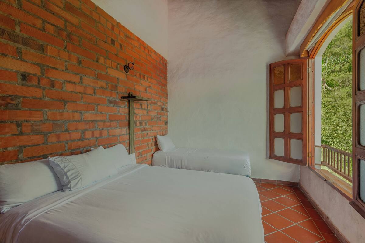 Quarto com duas camas, parede de tijolos, janela com moldura de madeira e vista para o verde lá fora.