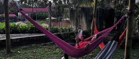 A hammock in a garden area with a fence, trees, and a building in the background.