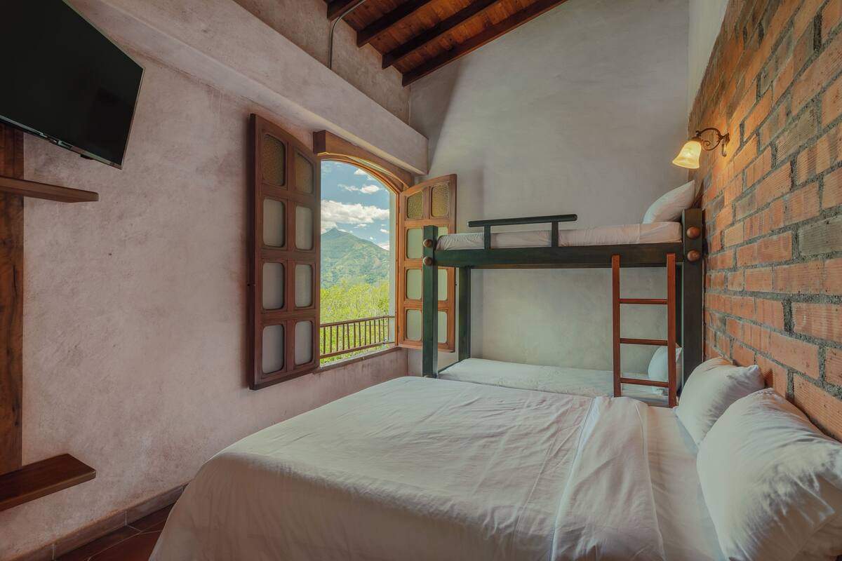 Quarto com beliche, televisão, janela com persiana aberta e parede de tijolos.