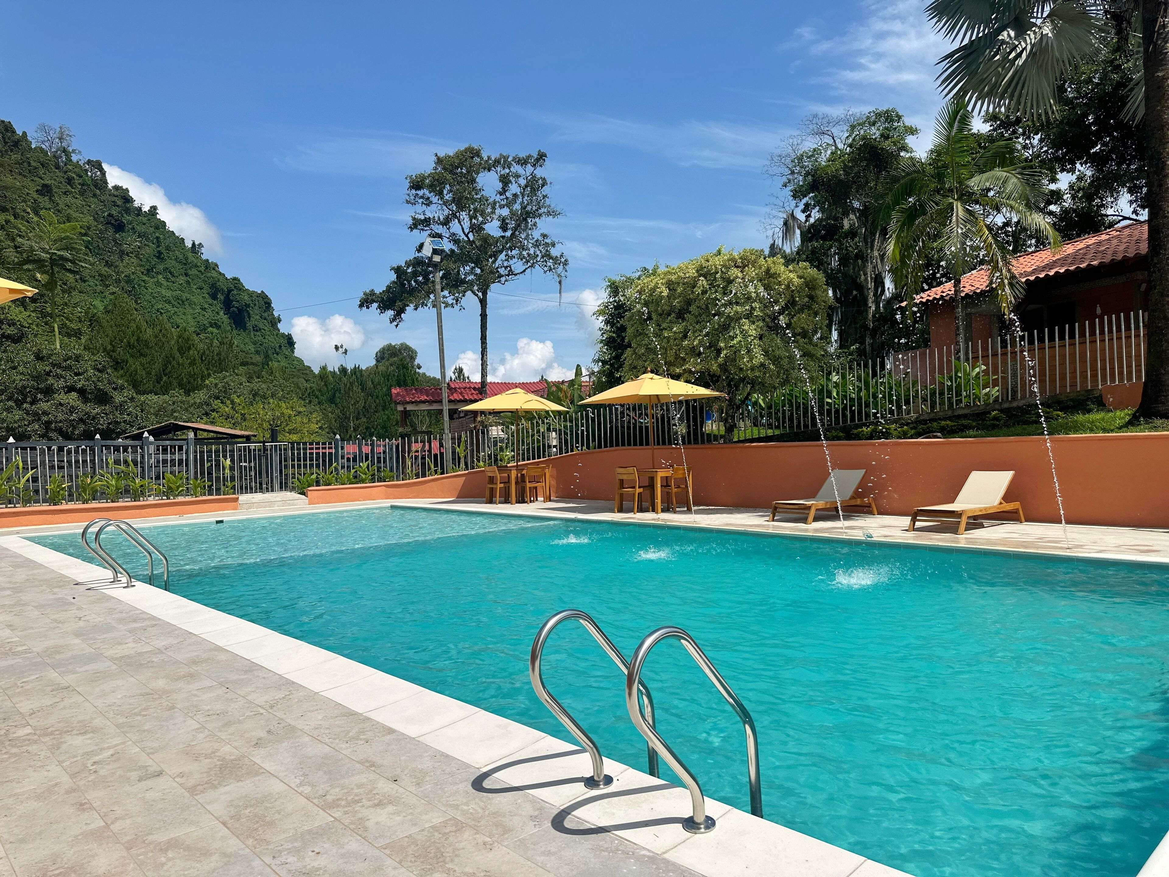 Une piscine à l’eau d’un bleu cristallin, entourée d’une terrasse carrelée. On y trouve des chaises longues et des parasols, et une clôture délimite l’espace.