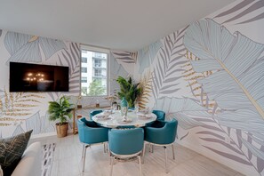 A dining area with a round table set for four, surrounded by teal chairs, a large potted plant, and a wall-mounted TV.