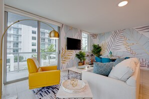 A modern living room with a yellow armchair, a white sofa, and a coffee table. There is a large window with a view of buildings and a flat-screen TV mounted on the wall.