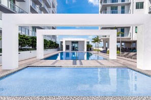 Un espace piscine moderne avec une piscine à l’eau d’un bleu limpide, des piliers blancs et une vue sur les bâtiments environnants.