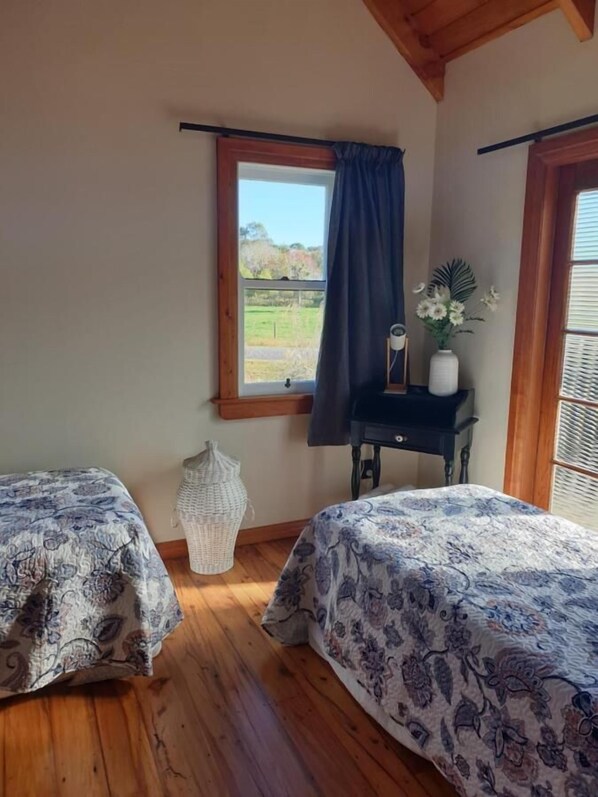 A bedroom with two beds, a window with a view of a field, a small table with a vase of flowers, and a wicker basket. - The Blackhouse Cottage (Ruatapu)