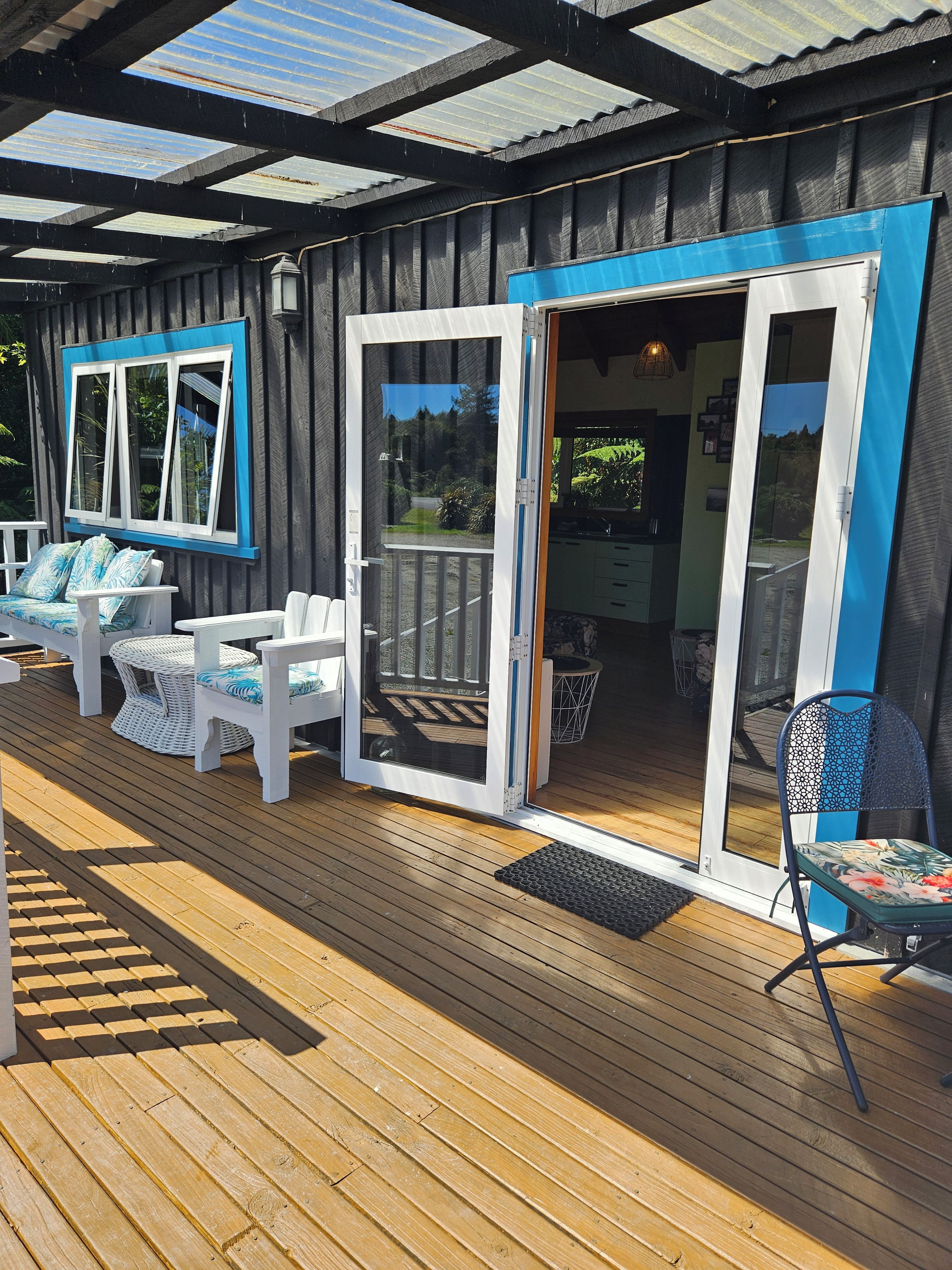 A covered outdoor deck with a wooden floor, white furniture, and a glass door leading to an interior space.
