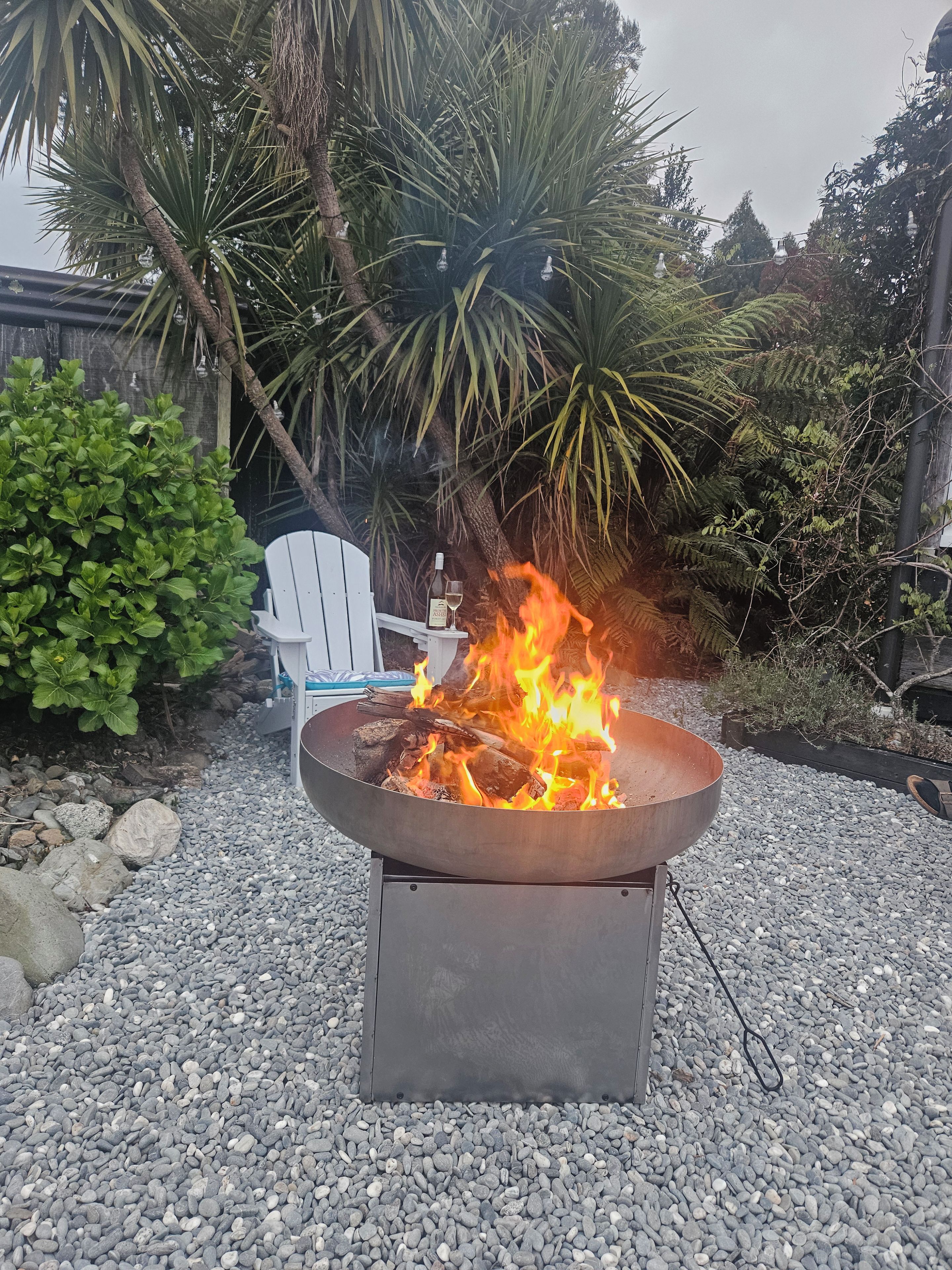Un hogar con leña, rodeado de guijarros, en un jardín con una silla y plantas.