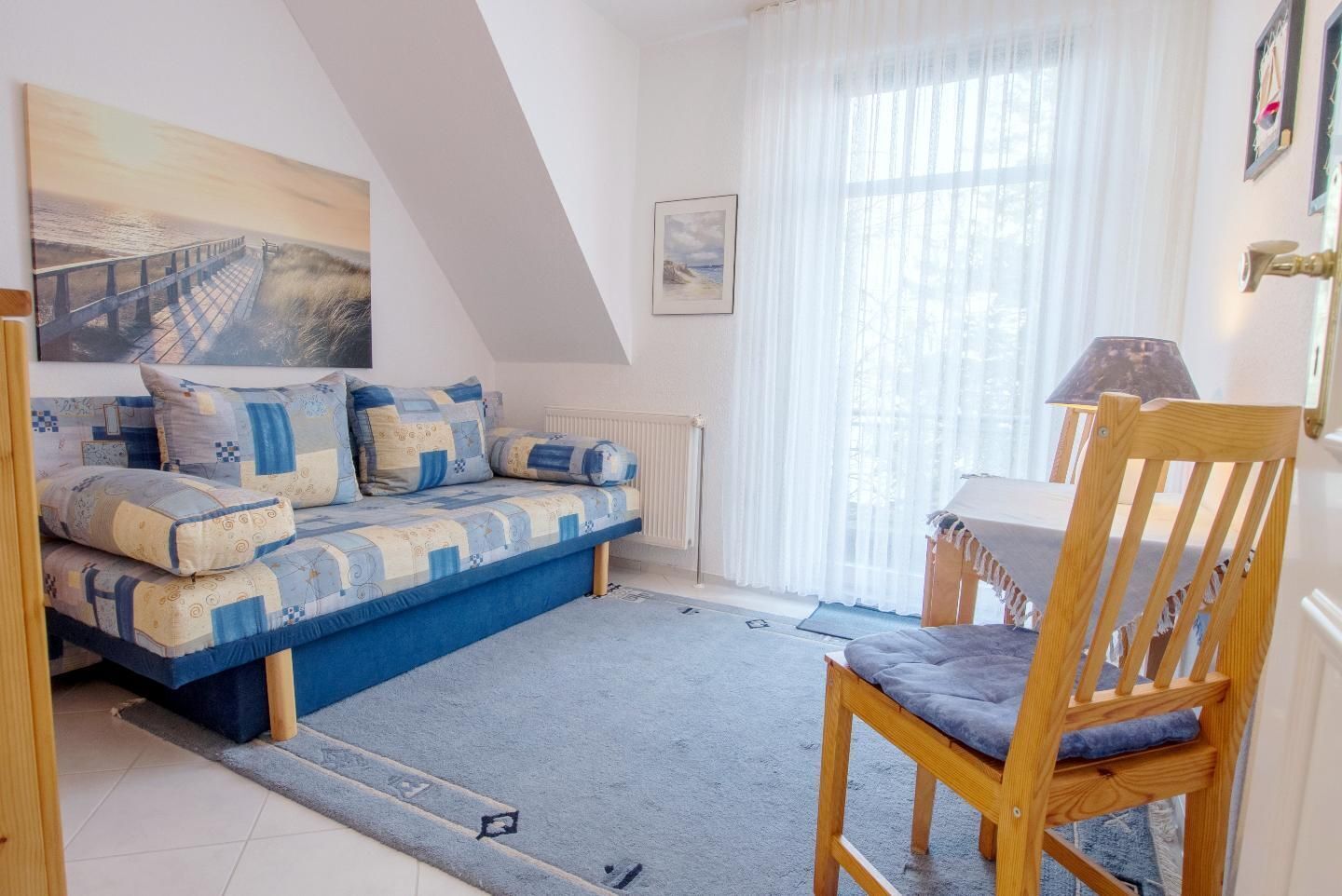 A cozy room with a blue sofa, a wooden chair, a table, and a painting of a pier.
