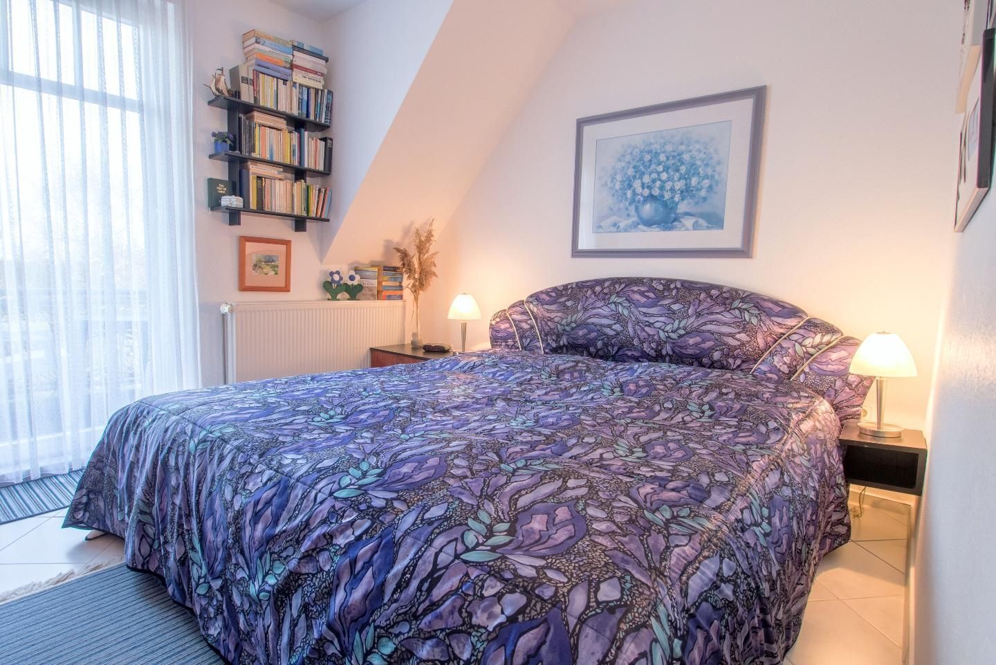 A bedroom with a large bed, a bookshelf, a framed picture, and a window with curtains.