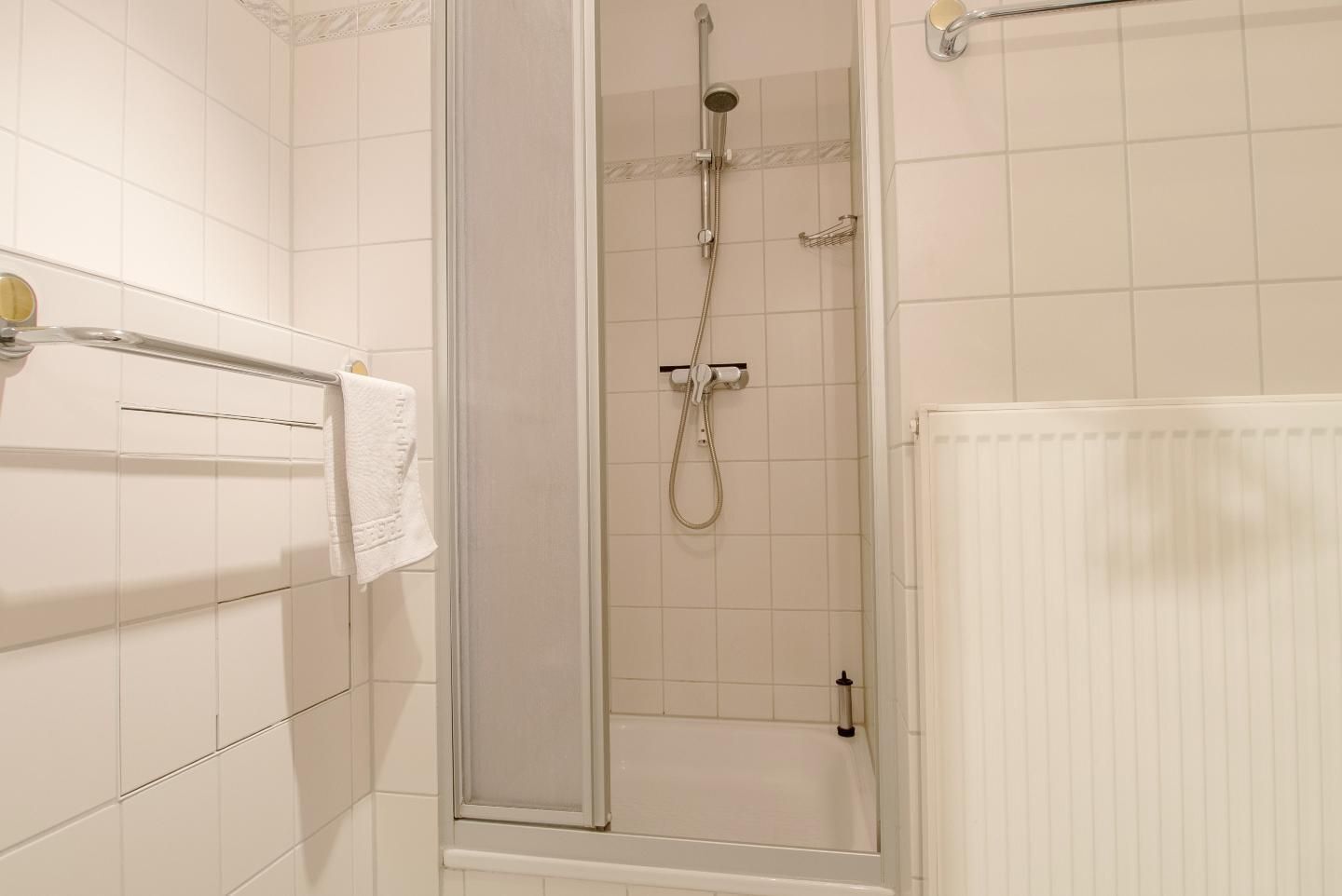 A bathroom with white tiled walls, a glass shower enclosure, a white radiator, and a towel rack with a white towel.