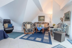 A cozy living room with a high ceiling, a leather armchair, a floral sofa set, a small table with a blue tablecloth, and a grey ottoman. - Apartment house Boddenblick "Himmelkieker" - Spacious apartment on the 2nd floor (Zingst)