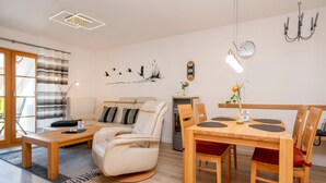 A modern living room with a sofa, a dining table with chairs, a fireplace, and a wall-mounted clock. - Terraced house with garden, terrace and heated pool in Zingst - vacation park (Zingst)