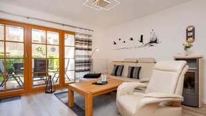A modern living room with a sofa, a coffee table, and a chair. There is a sliding glass door leading to an outdoor area with chairs. - Terraced house with garden, terrace and heated pool in Zingst - vacation park (Zingst)