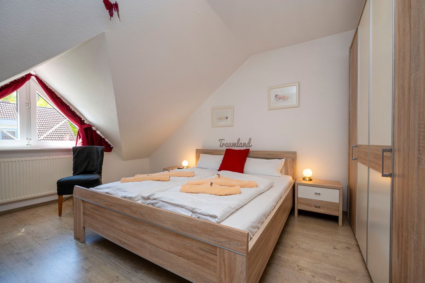 A bedroom with a sloped ceiling, a bed with white linens and a red pillow, a chair, a bedside table with a lamp, and a wardrobe.